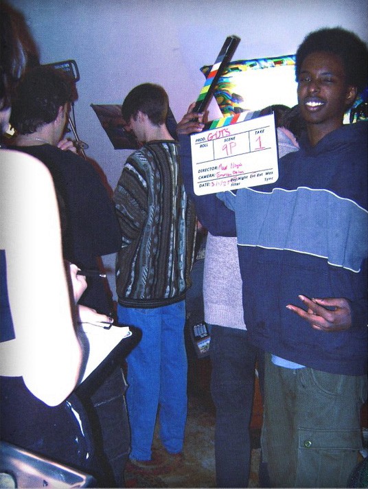 A smiling crew member holds a clapperboard beside the camera.